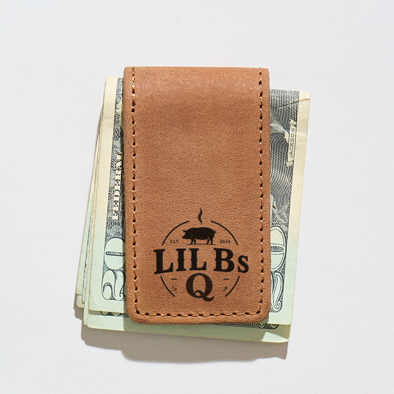 Brown leather money clip with 'LIL BS Q' branding clipped on a stack of US dollars.