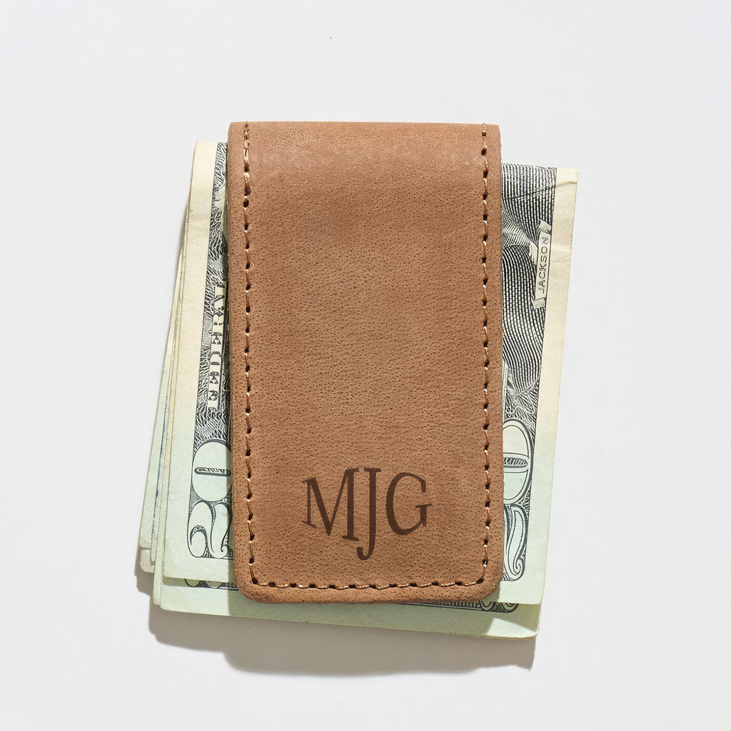 Brown leather money clip with 'MJG' monogram clipped onto a stack of cash against a white background